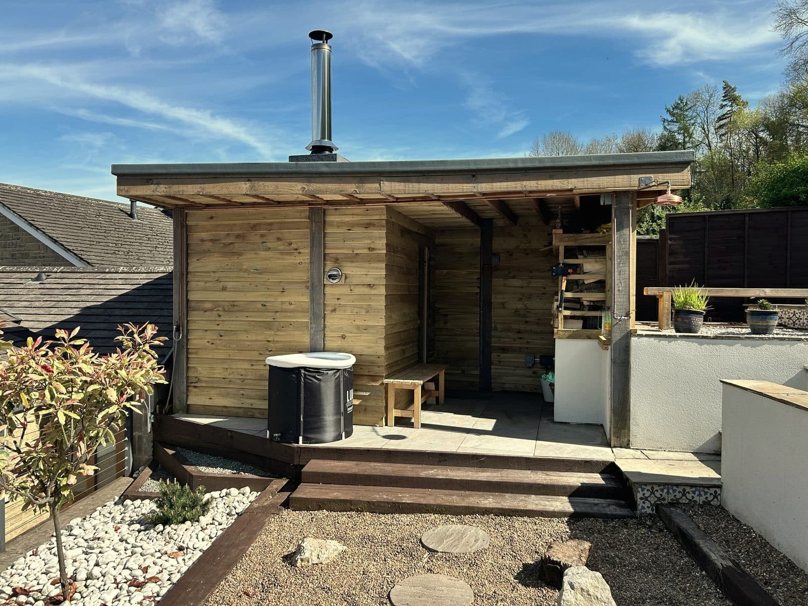 Finished gazebo sauna under a blue sky — flue up, log store at the covered end, Lumi plunge tub on the deck, sleeper steps below