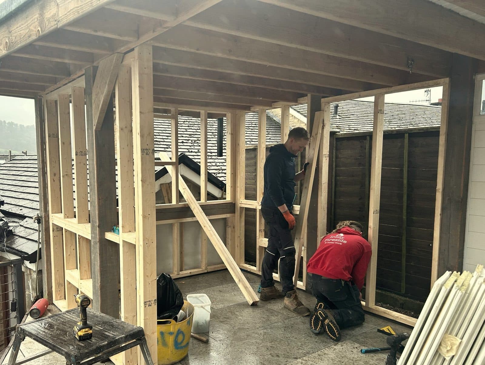 Timber frame of a garden sauna going up on a rainy day, two Irony Fireside builders at work with Peak District rooftops behind
