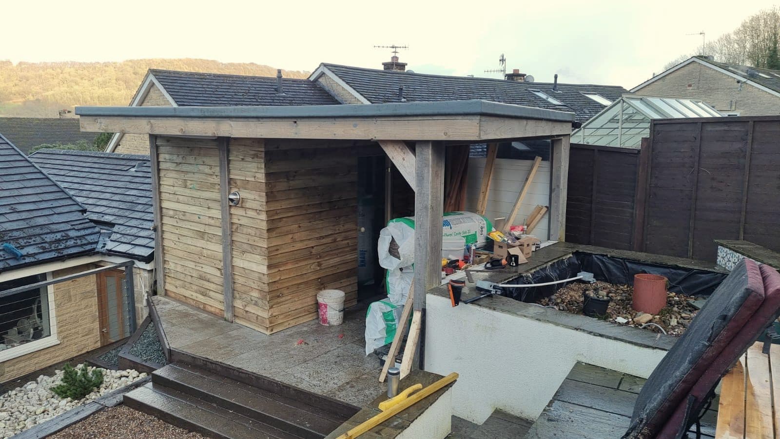 Gazebo mid-conversion on a wet day — new roof edge on, insulation bags staged, the old pond bed lined and empty beside it