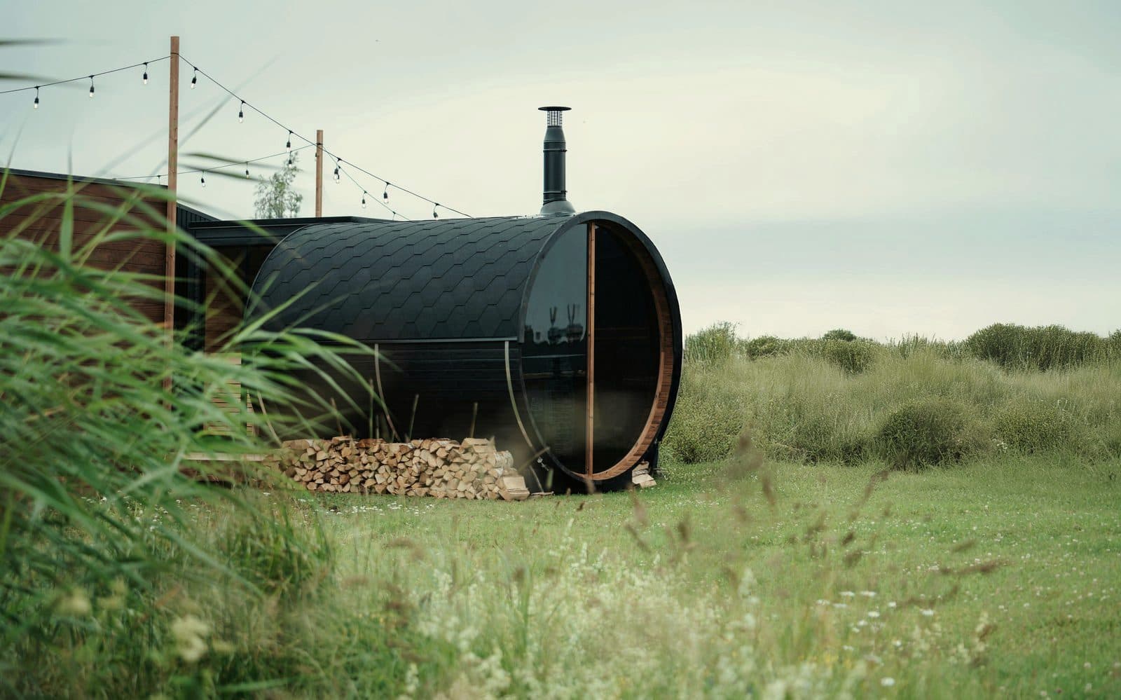 Black barrel sauna with chimney and stacked firewood in a rural meadow setting