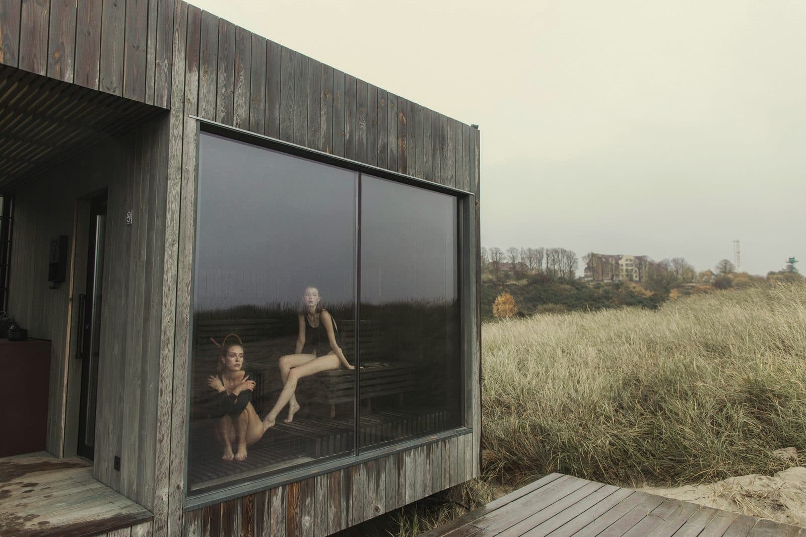 Timber-clad coastal sauna cabin with large glass window, dune grass and overcast sky