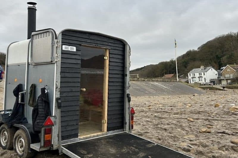 Black-clad horsebox sauna with chimney parked on a sandy beach with coastal village behind