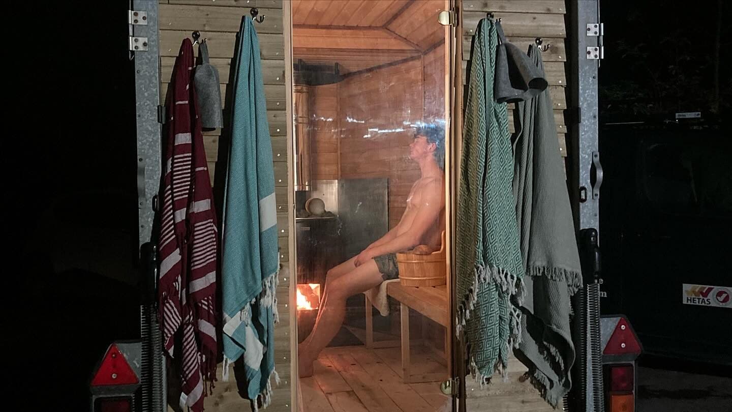 Person seated inside a horsebox sauna at night with wood-fired stove burning and towels hanging outside