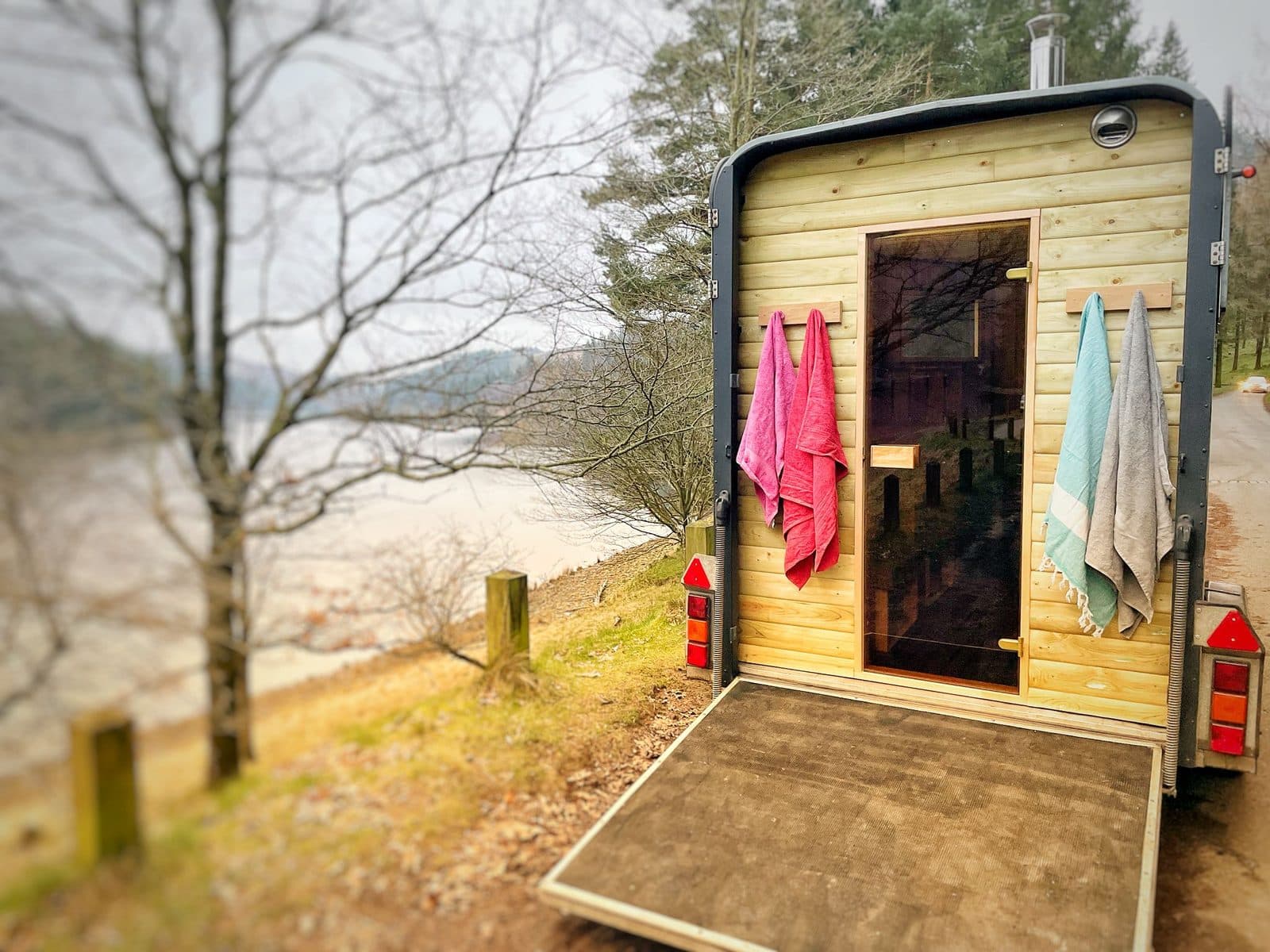 Timber-clad horsebox sauna with towels and ramp open overlooking a lake and hillside