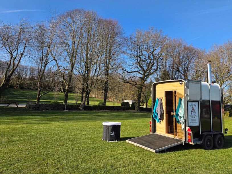 Horsebox sauna with cold plunge tub parked on green parkland under a clear blue sky
