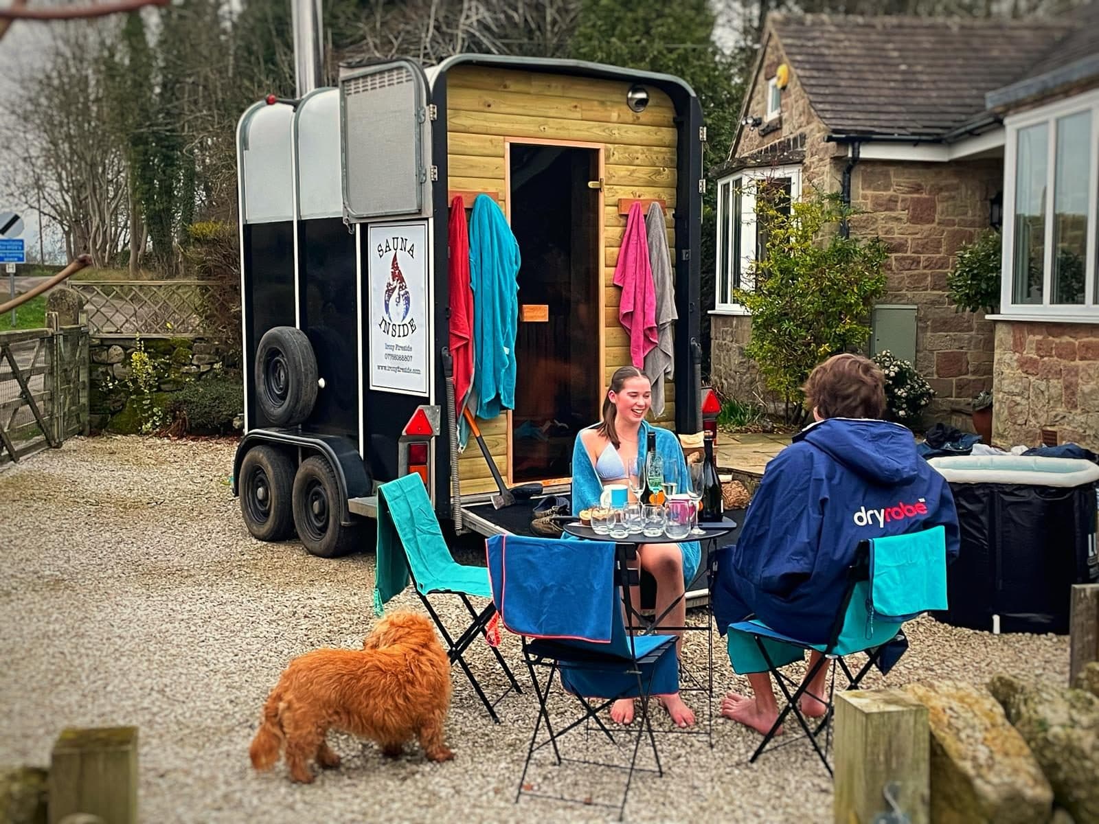 Two people relaxing at a table outside a horsebox sauna with drinks and a dog in a rural garden