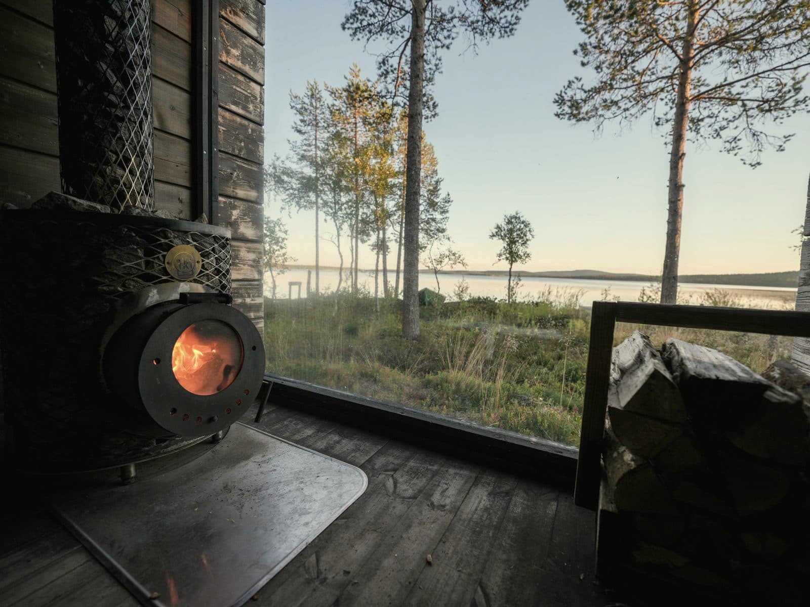 Wood-burning sauna stove with fire glowing on a lakeside deck at sunset with birch logs stacked nearby